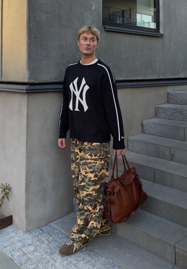 Homem jovem vestindo um suéter preto com um logotipo branco, calças camufladas, segurando uma grande mala de couro castanha, em pé ao lado de escadas e parede de cimento.