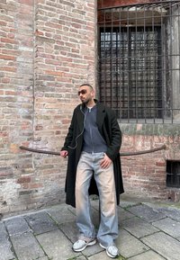 Homme portant des lunettes de soleil, un long manteau noir, un pull gris, un jean large clair et des baskets, debout devant un mur en briques avec une fenêtre grillagée à l'extérieur.