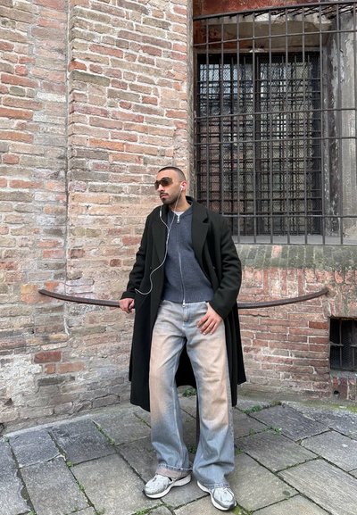 Hombre con gafas de sol, abrigo largo negro, suéter gris, jeans anchos claros y zapatillas de deporte, de pie contra una pared de ladrillos con una ventana con rejas al aire libre.