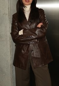 Γυναίκα με μακριά σκούρα μαλλιά, ντυμένη με καφέ δερμάτινο σακάκι πάνω από λευκό ζιβάγκο, με σταυρωμένα τα χέρια, στεκόμενη μπροστά σε τσιμεντένιους και μεταλλικούς τοίχους.