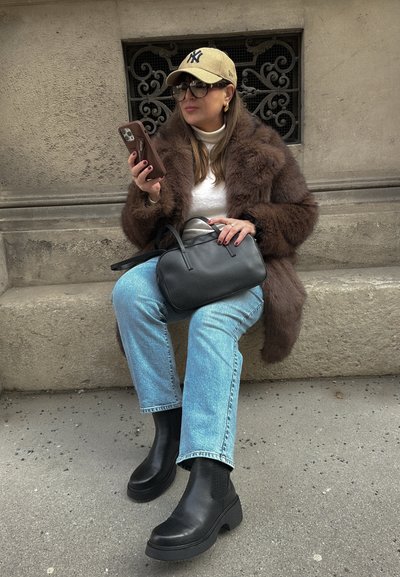 Abrigo de piel sintética marrón, camiseta blanca, jeans azul claro, botas chelsea negras, sosteniendo un teléfono inteligente, con un bolso negro en el regazo.