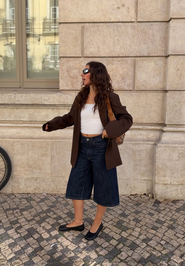 Une femme portant des lunettes de soleil, un blazer brun, un haut blanc, un pantalon jupe-culotte en jean et des chaussures plates gesticule dans une rue pavée, à côté d’un bâtiment en pierre.