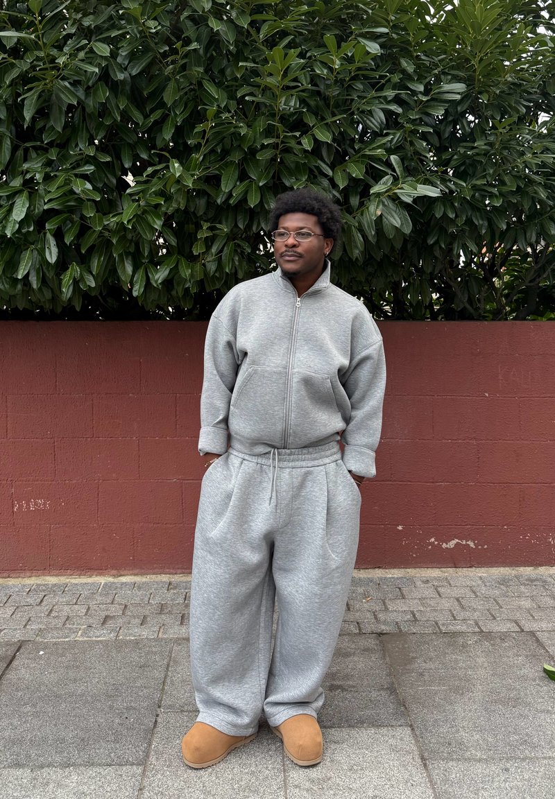 Felpa grigia con zip e pantaloni della tuta abbinati a vestibilità ampia. Pantofole marroni. Fogliame verde sullo sfondo, parete rossa con texture dietro.