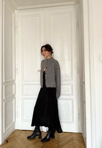 Cardigan gris en maille avec un col montant et une fermeture à boutons, porté sur une robe midi en satin noir et des bottines noires, devant un mur blanc.