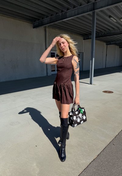 Joven mujer con tatuajes que lleva un vestido marrón sin mangas y botas negras hasta la rodilla, sosteniendo un bolso con estampado en blanco y negro al aire libre.
