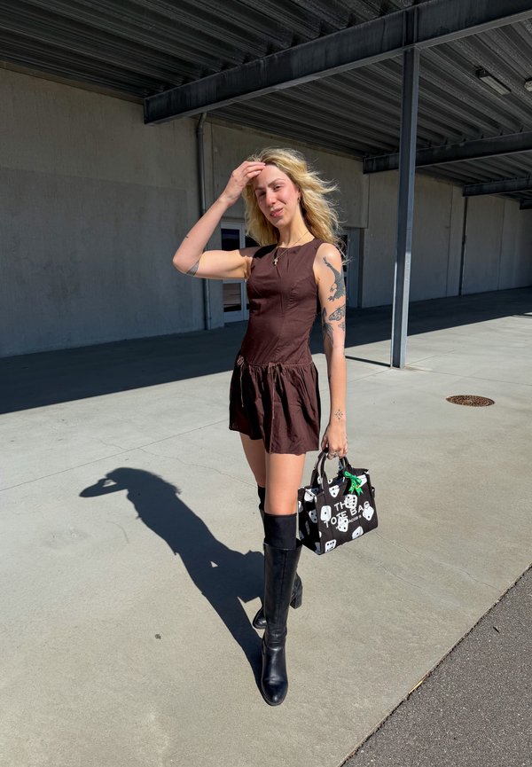 Mladá žena s tetovaniami oblečená v hnedých šatách bez rukávov a čiernych čižmách po kolená drží vonku čierno-bielu vzorovanú kabelku.