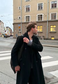 Crni kaput s dvostrukim kopčanjem i sivim manšetama, nošen preko svijetle majice. Tamna torba prebačena preko ramena, stoji na pješačkom prijelazu blizu zgrada.