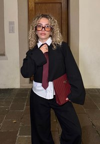 Czarna, oversizowa kurtka na białej koszuli, bordowy krawat i szerokie czarne spodnie. Trzyma czerwony, żeberkowany portfel z złotym okuciem. Kręcone włosy.