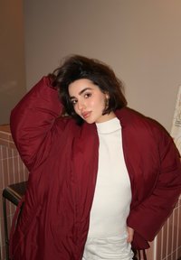 Burgundská péřová bunda s širokými rukávy přes bílý rolák. Tkanina je lesklá, s viditelným švem a kapsami.