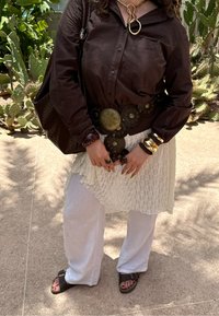 Camisa castanha de botões, blusa branca em camadas, calças largas brancas. Cinto circular grande com acentos metálicos, pulseiras robustas e uma bolsa de palha.