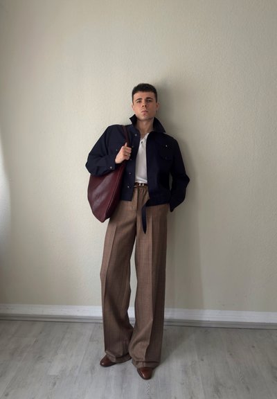 Joven de pie en el interior con una chaqueta azul marino, camisa blanca, pantalones de pierna ancha marrones con cuadros, zapatos marrones y lleva un gran bolso de cuero burdeos.