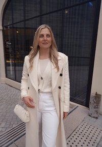 Casaco longo branco com botões pretos, suéter creme e calças brancas. Asegurando uma pequena bolsa bege, de pé sobre um pavimento de pedras padrões.