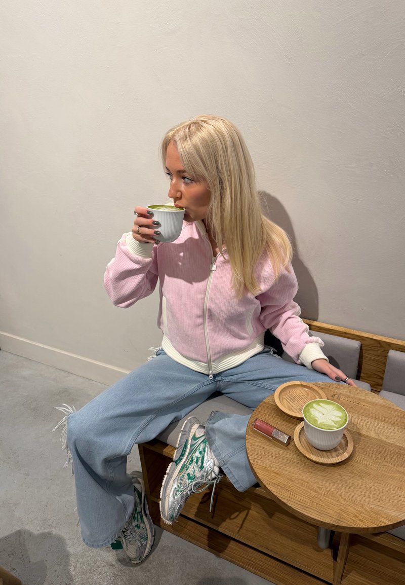 Femme en veste rose et jean assise sur un banc, buvant du matcha dans une tasse blanche, avec un autre matcha et un rouge à lèvres posés sur une table en bois.