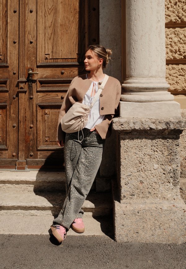Cárdigan corto beige, top blanco con lazo, pantalones gris con estampado y zapatillas rosas. Bolso beige con detalle de cordón. Fondo de pared de piedra.