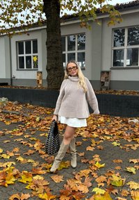 Gestreepte beige en roomkleurige coltrui, witte jurk, beige kniehoge laarzen en zwarte handtas met studs, te midden van herfstbladeren.