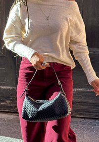 Zwarte geweven handtas met een gebogen ontwerp en riem, gecombineerd met een gebroken witte gebreide trui en bordeauxrode broek, met zichtbare gebreide textuur.