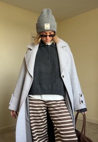 Grey wool blend overcoat, dark jumper, white shirt, striped brown and cream trousers, grey knitted beanie, and dark sunglasses. Brown handbag.