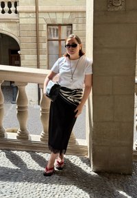 Gonna nera con bordi in pizzo, t-shirt bianca, cintura con stampa zebra e borsa nera. Indossa ciabatte rosse e occhiali da sole. Sfondo con un pilastro in pietra.