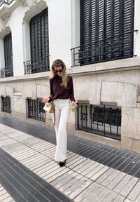 Pantaloni bianchi a zampa d'elefante, maglietta scura a maniche lunghe, occhiali da sole neri e con una bevanda in mano. Porta una piccola borsa a tracolla color crema. Sfondo di marciapiede e edificio.