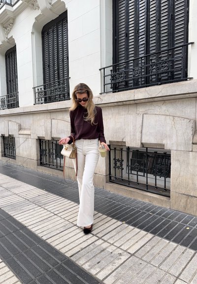 Pantalones acampanados blancos, top de manga larga oscuro, gafas de sol negras y sosteniendo una bebida. Lleva un pequeño bolso de crema. Fondo de acera y edificio.