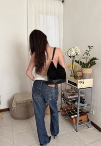 Femme aux longs cheveux vêtue d'un jean et d'un haut dos nu, portant un sac à bandoulière noir, debout dans une pièce avec des plantes et une petite table de baby-foot.