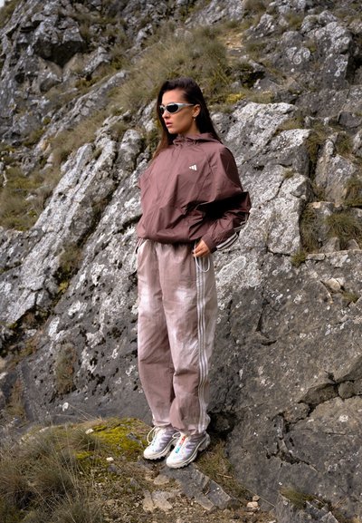 Mujer con chaqueta marrón y pantalones de chándal beiges, usando gafas de sol, se encuentra en una ladera rocosa con escasa vegetación.