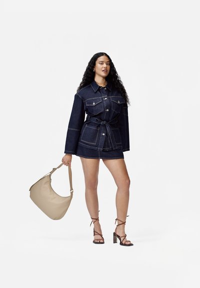 Vestido de chaqueta de mezclilla en azul oscuro con cintura ceñida, que presenta detalles de bolsillos y botones plateados. Sosteniendo un bolso de hombro beige.