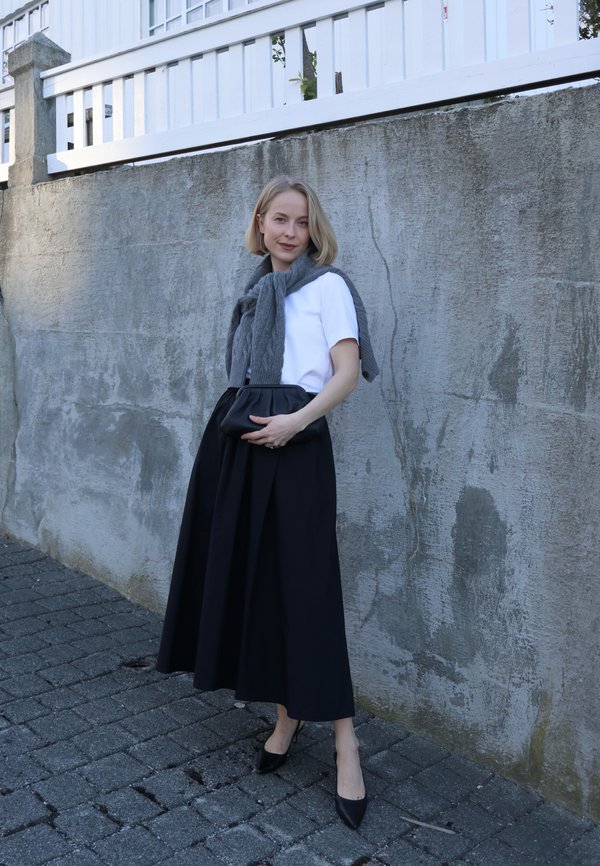 Femme debout sur un pavé en pierre devant un mur en béton, portant un haut blanc, une jupe noire, un pull gris posé sur les épaules, tenant une pochette noire.
