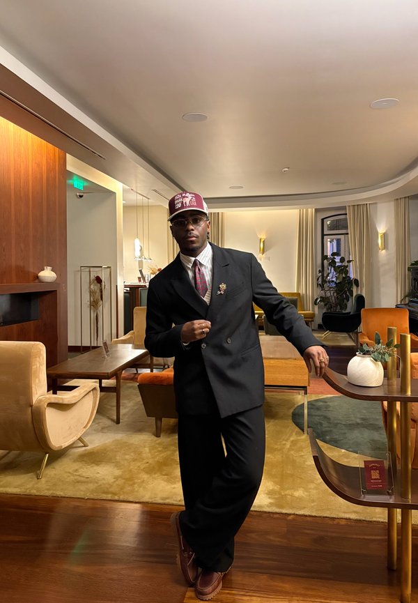 Hombre con traje negro de doble botonadura, corbata estampada y gorra granate, de pie en una sala de estar elegante con muebles de mediados de siglo y una iluminación cálida.