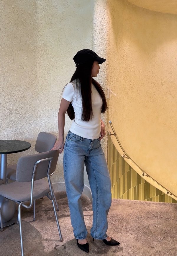 Une femme portant une casquette noire, une chemise blanche et un jean se tient à côté de chaises et d’une rampe d’escalier incurvée.