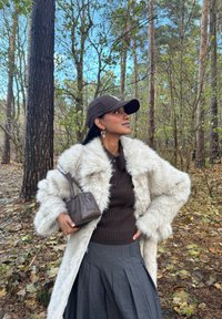 Vrouw draagt een beige nep-bontjas, bruine trui, geplooide grijze rok en een bruine pet, houdt een bruine handtas vast en staat in een bos met gevallen bladeren.