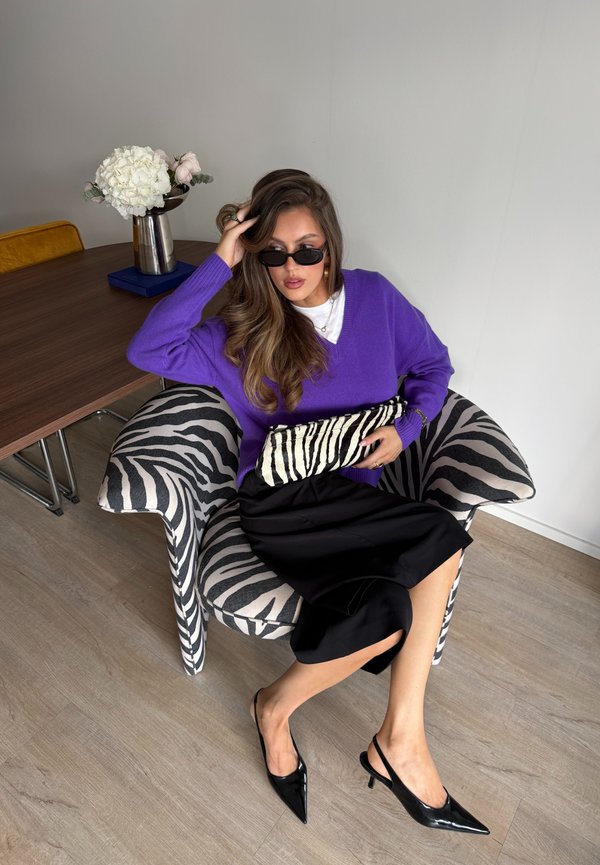 Femme en pull violet et jupe noire assise sur une chaise à imprimé zèbre tenant une pochette zébrée avec des fleurs sur la table à proximité.
