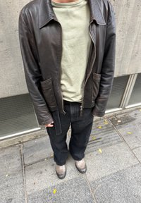 Chaqueta de cuero marrón con cierre de cremallera, camiseta verde oliva debajo, pantalones negros de pierna ancha y botas grises, vistos sobre un fondo de hormigón.