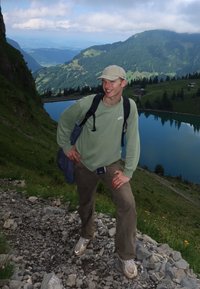 Mladý muž v kšiltovce a s batohem stojí na kamenité stezce v horách s výhledem na jezero a lesnaté kopce pod zataženou oblohou.