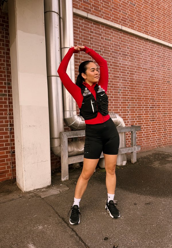 Une femme en tenue de course s’étire, les bras au-dessus de la tête, près d’un mur en briques. Elle porte un gilet d’hydratation et des chaussures de course.