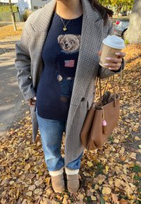 Mētelis ar haizivs zobu rakstu, tumši zils adīts džemperis ar lāča grafiku, zili džinsi, brūnas zamša zābaki ar baltu oderējumu, brūna soma ar bārkstīm.