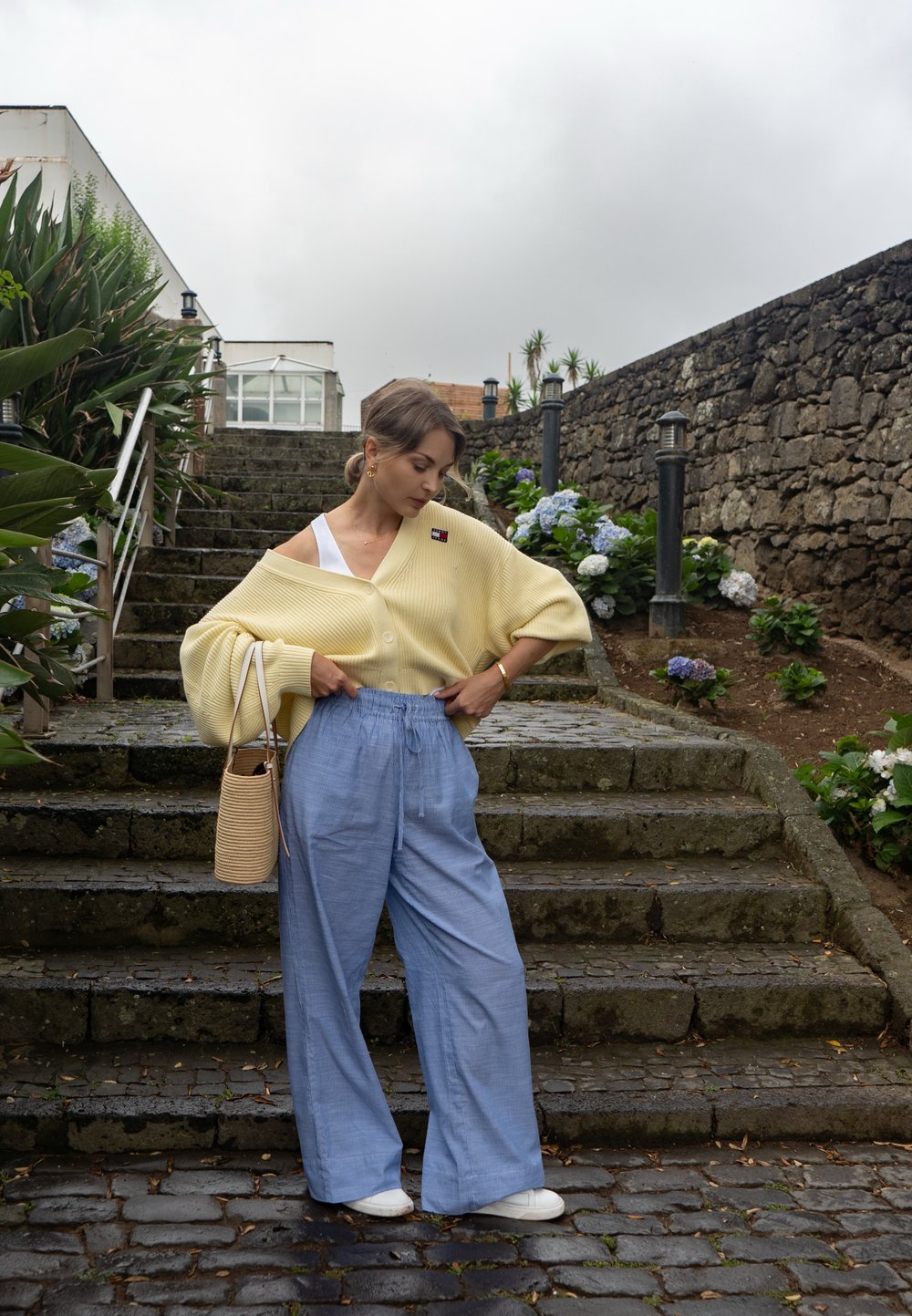 Suéter amarelo com design ombro-a-ombro sobre uma blusa branca, combinado com calças largas azul-claro. Inclui uma bolsa de palha.