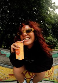 Óculos de sol com uma armação verde-clara, vestindo um moletom preto, segurando um cartão de sumo de laranja. O fundo apresenta árvores e graffiti.