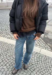 Veste matelassée noire, pull marron, jean large bleu clair, et bottines marron. Fond en pavés texturés.