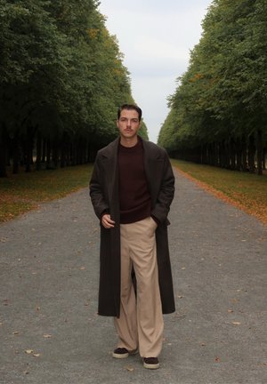 Langer dunkler Mantel über einem kastanienbraunen Pullover, kombiniert mit weiten beigen Hosen und braunen Turnschuhen, vor einem von Bäumen gesäumten Weg mit gefallenem Laub.