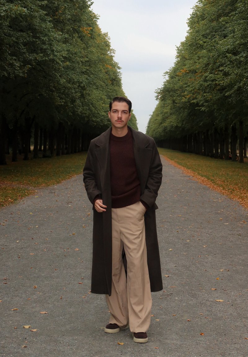 Long manteau sombre sur un pull bordeaux, associé à un large pantalon beige et des baskets brunes, sur un chemin bordé d'arbres avec des feuilles tombées.