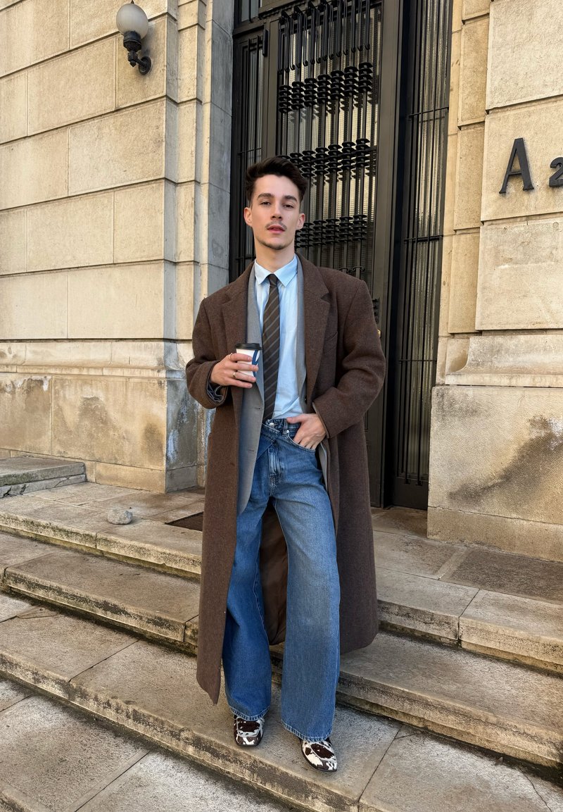 Νεαρός άνδρας που φοράει μακρύ καφέ παλτό, μπλε πουκάμισο, γραβάτα με ρίγες, φαρδιά τζιν και παπούτσια με μοτίβο αγελάδας, κρατώντας ένα φλιτζάνι καφέ σε πέτρινα σκαλιά.