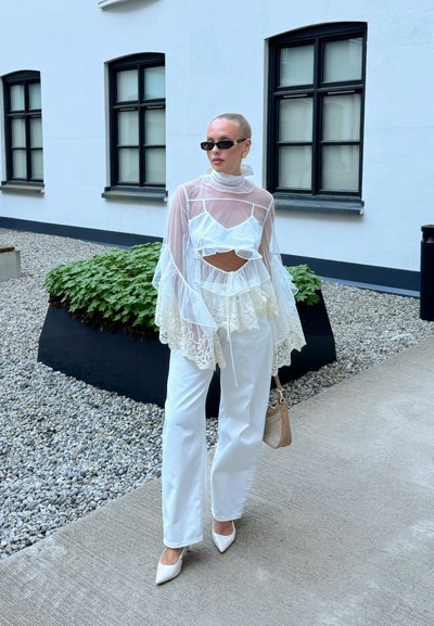 Mujer con moño rubio que lleva una blusa blanca de encaje transparente, pantalones blancos, tacones blancos, gafas de sol negras y sostiene un bolso beige al aire libre junto a un edificio.