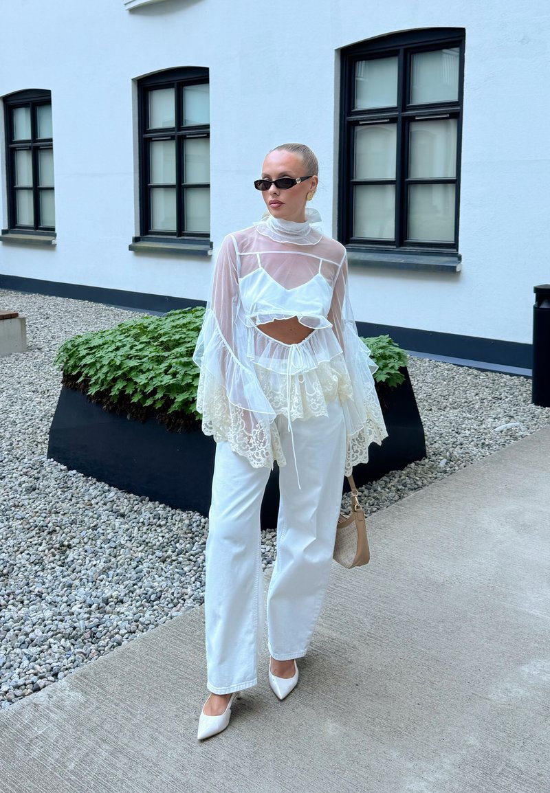 Femme avec un chignon blond portant un chemisier en dentelle blanche transparente, un pantalon blanc, des talons blancs, des lunettes de soleil noires et tenant un sac à main beige à l'extérieur près d'un bâtiment.