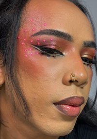 Primer plano de un rostro con pedrería rosa en la frente, piercing dorado en el septum y maquillaje audaz con tonos tierra y delineado preciso.