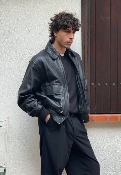 Chaqueta bomber de cuero negro con cuello camisero, cierre de cremallera y dos bolsillos delanteros, combinada con un suéter oscuro y pantalones negros sueltos.