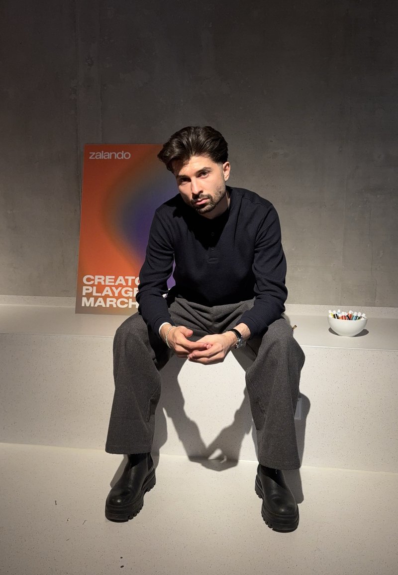Homme aux cheveux foncés et à la barbe assis sur un rebord portant un pull noir, pantalon gris et bottes noires ; affiche colorée Zalando et bol avec des marqueurs à proximité.