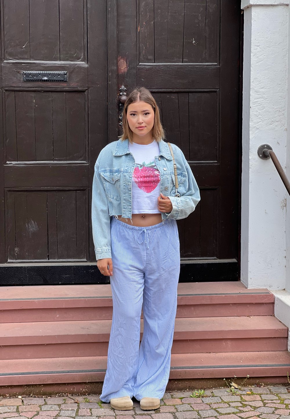 Izgriezta džinsu jaka, balts T-krekls ar zemeņu grafiku un gaiši zili svītraini platpantus. Stāv uz pakāpieniem blakus tumšiem koka durvīm.