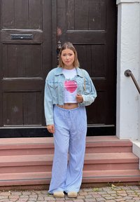 Izgriezta džinsu jaka, balts T-krekls ar zemeņu grafiku un gaiši zili svītraini platpantus. Stāv uz pakāpieniem blakus tumšiem koka durvīm.