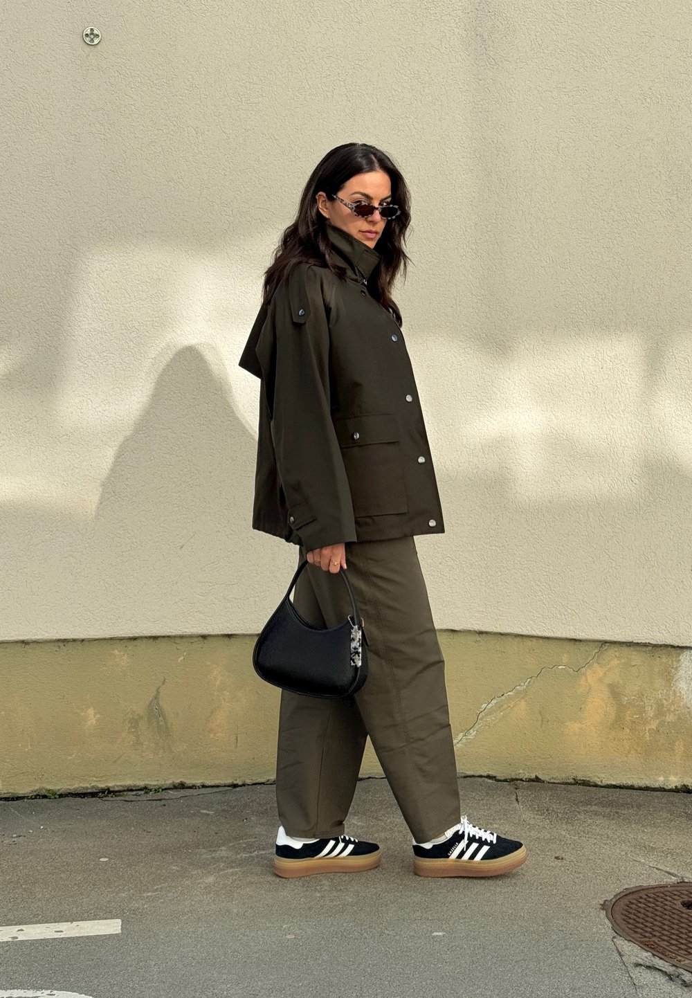 Ελαιόχρωμο αδιάβροχο μπουφάν με κουμπιά αρμάτωσης, συνδυασμένο με ελαιόχρωμες παντελόνες. Μαύρη τσάντα ώμου και μαύρα αθλητικά παπούτσια με λευκές ρίγες.